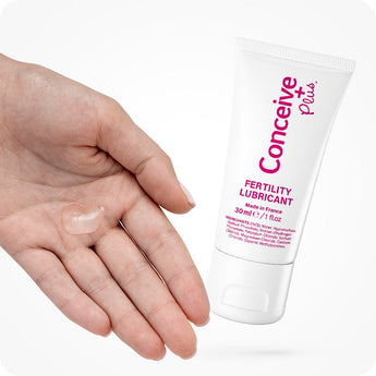Conceive Plus Fertility Lubricant (1fl.oz/30ml Tube) shown beside an open hand with a visible drop of clear gel, demonstrating product texture and ease of application for couples trying to conceive.